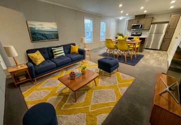 Modern open-plan living room and kitchen with a navy sofa, yellow pillows, wooden coffee table, yellow patterned rug, dining table with yellow chairs, and stainless steel appliances. The space is brightly lit and tidy.