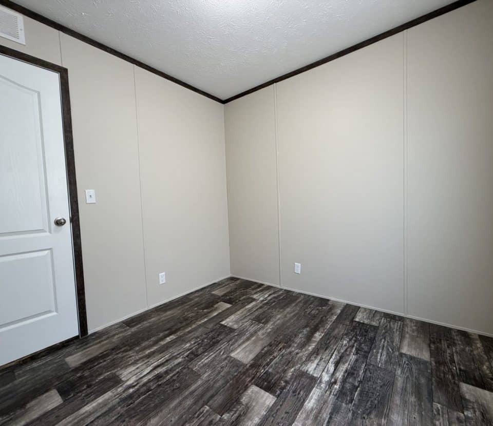 An empty room with beige walls, a white door, a white ceiling, dark trim, and wood-patterned vinyl flooring. There are electrical outlets on the walls and no furniture or decorations.
