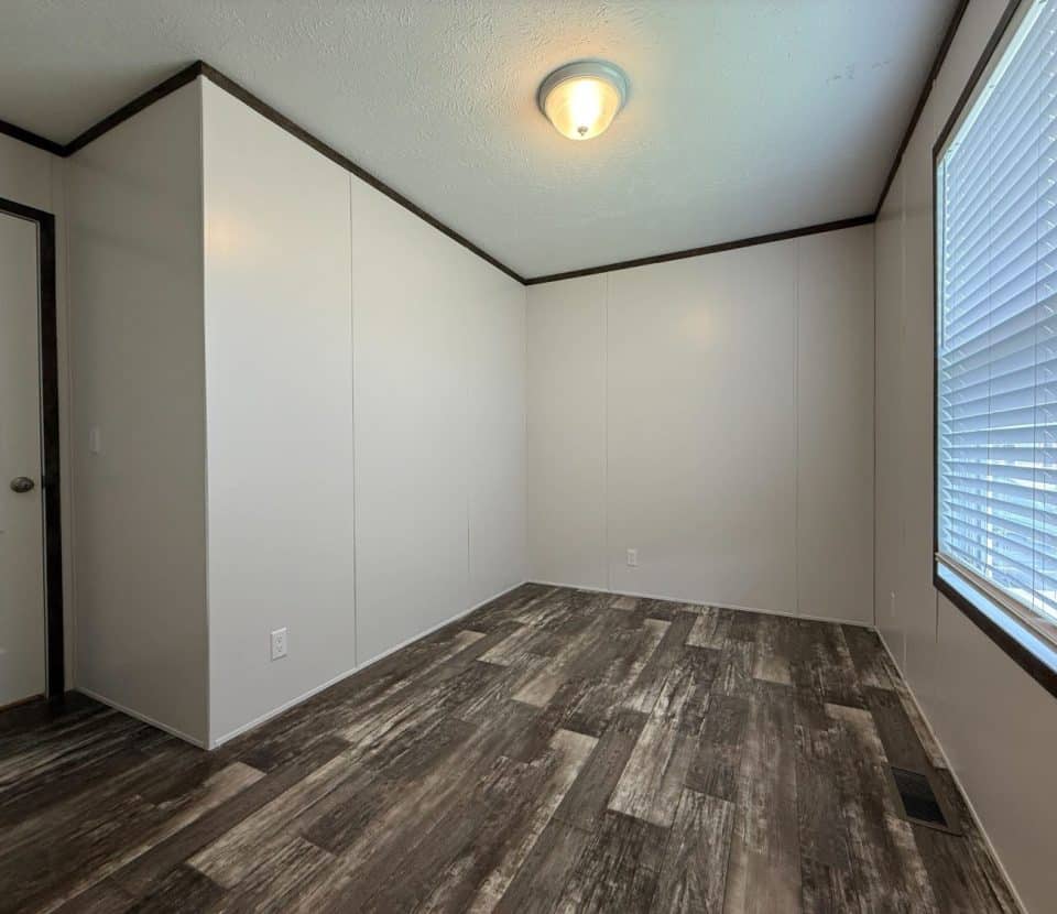 Empty room with light-colored walls, wood-patterned flooring, a closed white door, a large window with blinds, and a ceiling light fixture. The room is well-lit and unfurnished.