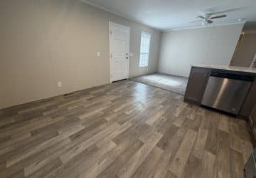 An empty room with wood-patterned vinyl flooring, beige walls, a white front door, a window, and a ceiling fan. Part of a kitchen counter with a built-in dishwasher is visible on the right.