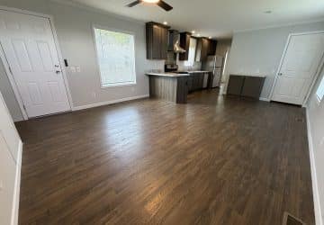 Open-concept living area with wood flooring, a ceiling fan, and large windows. The kitchen features dark cabinets, stainless steel appliances, and an island. White doors and neutral walls complete the modern space.