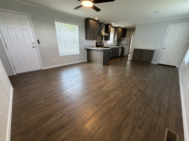 Open-concept living area with wood flooring, a ceiling fan, and large windows. The kitchen features dark cabinets, stainless steel appliances, and an island. White doors and neutral walls complete the modern space.