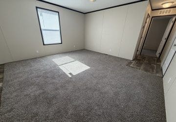 A bright, empty room with beige walls, gray carpet flooring, a window letting in sunlight, and a doorway leading to a hallway with wood-patterned flooring.