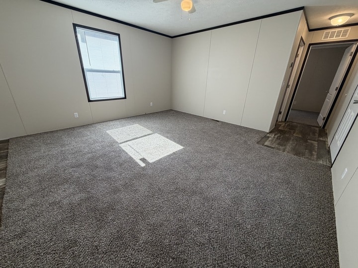 A bright, empty room with beige walls, gray carpet flooring, a window letting in sunlight, and a doorway leading to a hallway with wood-patterned flooring.