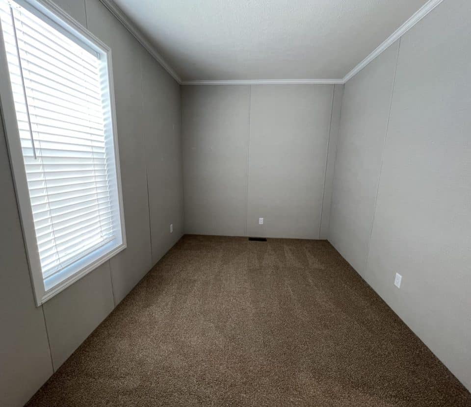An empty room with beige carpet, light-colored walls, a white ceiling, and a large window with closed blinds letting in natural light. There are electrical outlets on the walls.