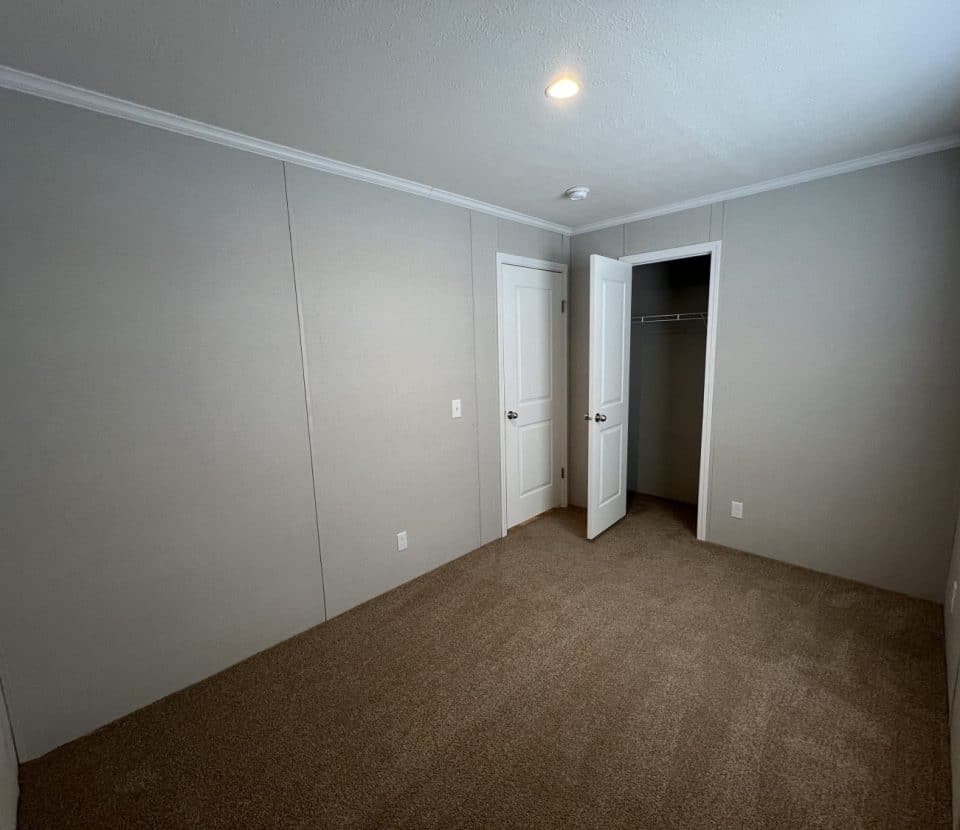 Empty carpeted room with beige walls, a ceiling light, and a window on the right. There are two white doors: one open to a closet with a hanging rod, and one leading out of the room.