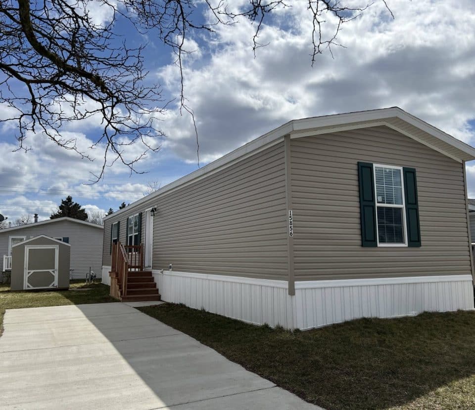 A beige manufactured home with green shutters sits next to a driveway and a small shed, under a partly cloudy sky with leafless tree branches in the foreground.