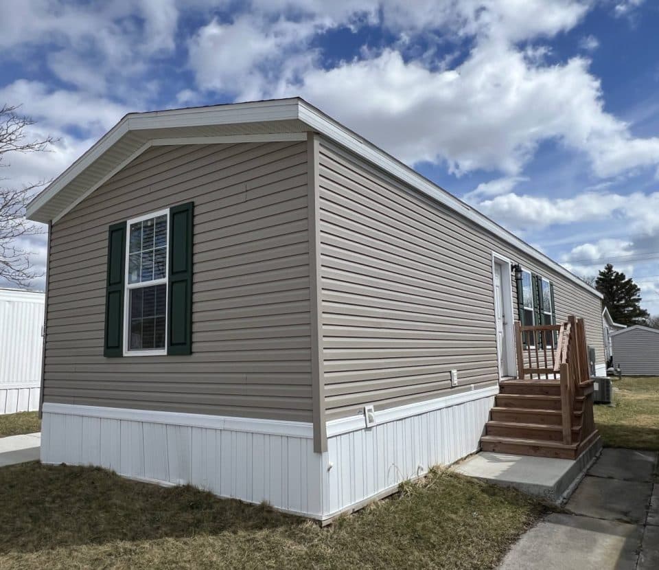 A beige mobile home with green shutters sits on a grassy lot. Wooden steps lead up to the entrance, and a concrete path runs alongside. The sky is partly cloudy.