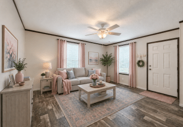 A bright living room with a gray sofa, pink and gray cushions, a coffee table with flowers, two windows with pink curtains, a ceiling fan, and decor in soft, neutral colors. A door and entryway are visible on the right.