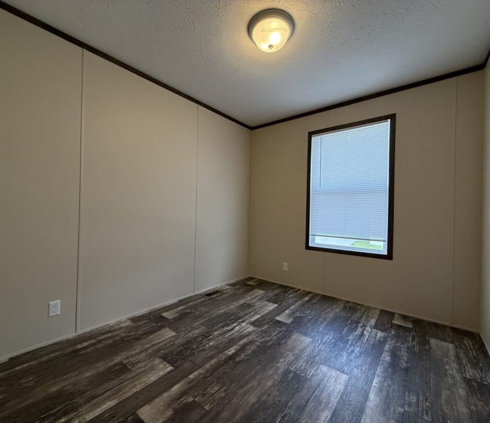 An empty room with beige walls, dark wood-style flooring, a single window with closed blinds, a ceiling light fixture, and a door partially visible on the left.