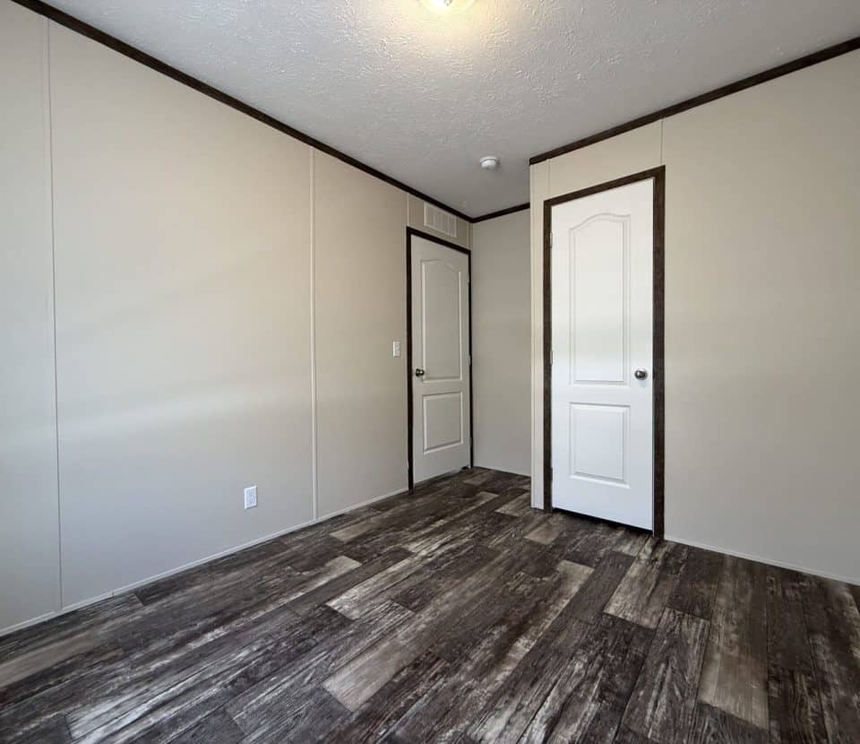 Empty room with beige walls, a white ceiling with a single light fixture, dark trim, and two white doors. The floor has a wood-like pattern in shades of brown and gray.