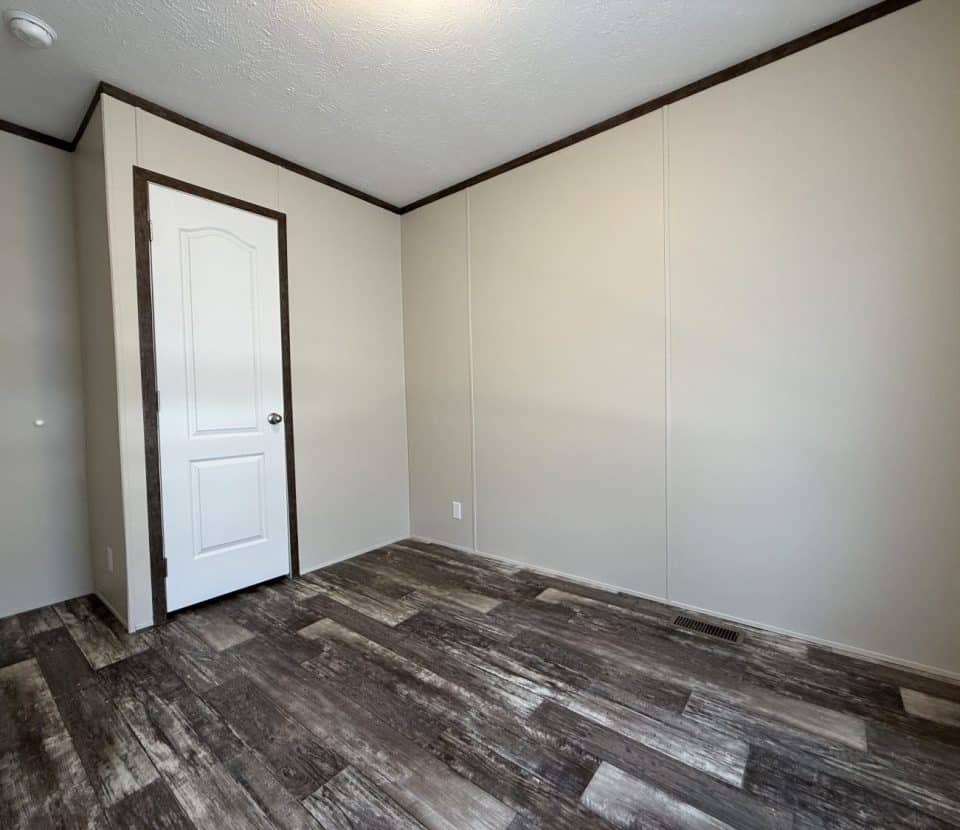 Empty room with beige walls, a white door with dark trim, and wood-patterned vinyl flooring. The room is well-lit and has a simple, clean appearance.