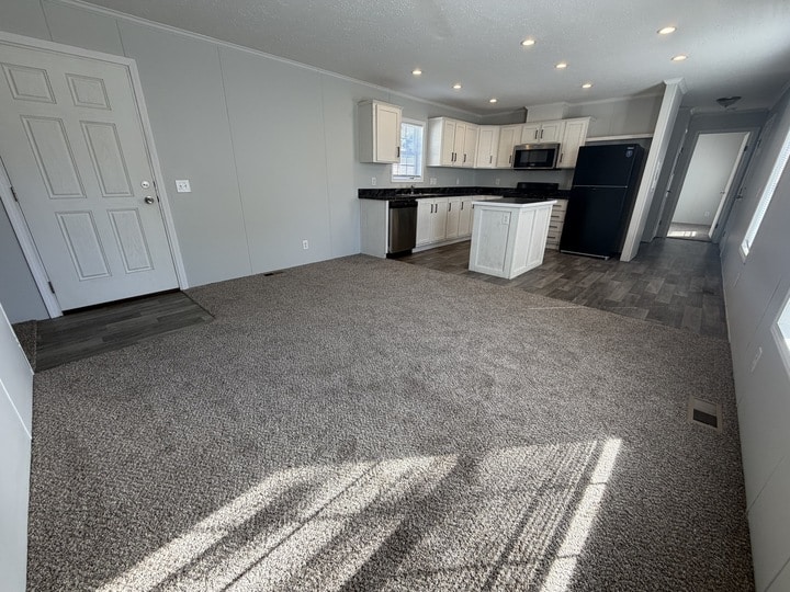 A bright, empty open-concept living area with carpeted flooring, a modern kitchen featuring white cabinets, black countertops, stainless steel appliances, and a small center island. Light streams in from unseen windows.