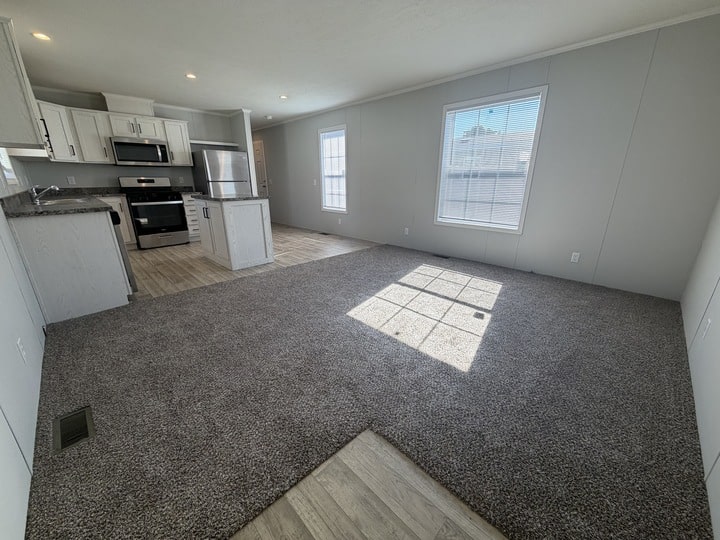 A bright, modern open-plan living area with gray carpet, large windows, and an adjacent kitchen featuring white cabinets, stainless steel appliances, and wood-style flooring. Sunlight streams in through the windows.