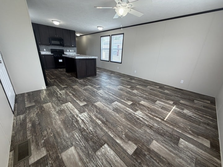 A modern open-concept kitchen and living area with wood-look vinyl flooring, neutral beige walls, dark cabinetry, and a ceiling fan. Large windows provide natural light. The space is empty and unfurnished.