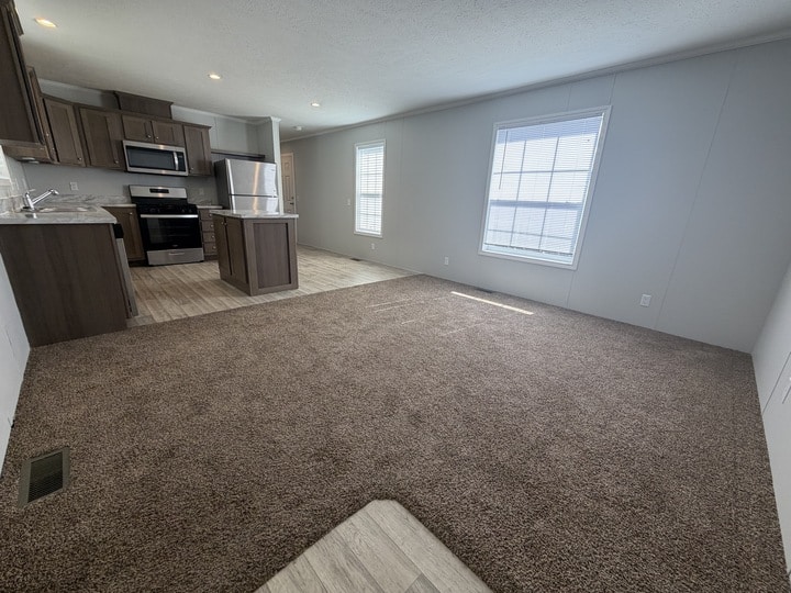 Open living area with brown carpet and a modern kitchen featuring stainless steel appliances, dark cabinets, and light tile flooring. Two large windows let in natural light. Walls are light-colored.