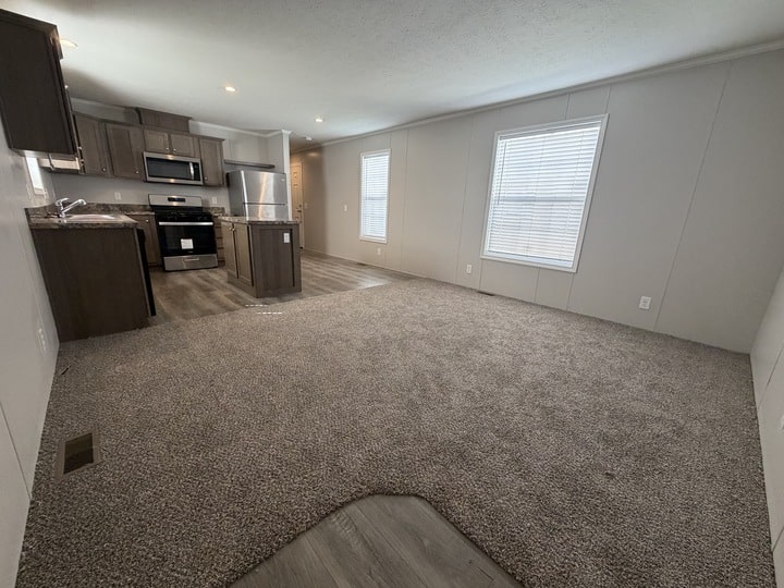 Open-concept living area with gray carpet, leading to a kitchen with dark cabinets, stainless steel appliances, and wood-look flooring. Large windows provide natural light throughout the space.