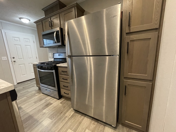 A modern kitchen with a stainless steel refrigerator, a gas stove with oven, a microwave above it, wooden cabinets, and wood-look vinyl flooring. A white door with a window is visible in the background.