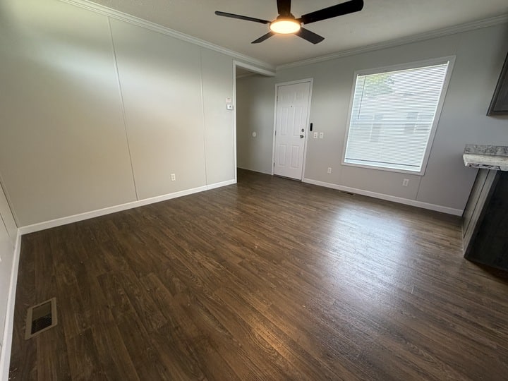 Empty living room with dark wood flooring, light gray walls, a large window, white front door, ceiling fan with light, and part of a kitchen counter visible on the right. Natural light fills the space.