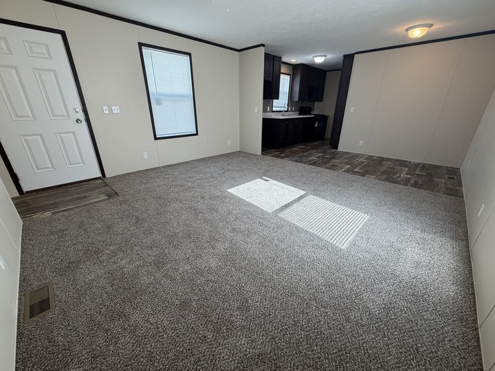 Spacious, unfurnished living room with gray carpet, beige walls, and a window letting in sunlight. A white door is on the left, and a kitchen area with dark cabinets and tile flooring is visible in the background.