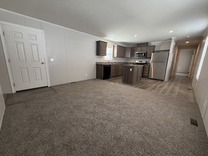 A spacious open-plan living area with carpeted floors, a modern kitchen featuring stainless steel appliances, and neutral-colored walls. A front door is on the left, and a hallway leads to other rooms.