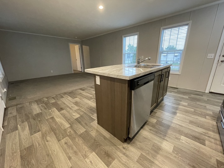 Modern kitchen with an island featuring a sink and dishwasher, adjacent to a living area with carpet, two large windows, and neutral-colored walls and floors.