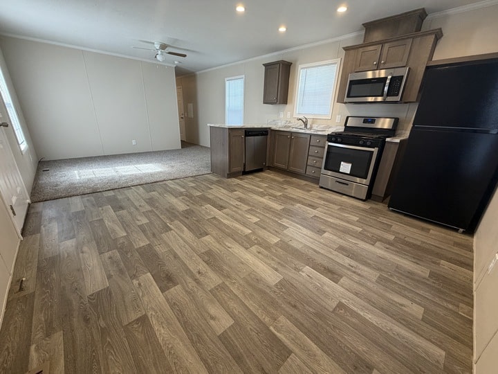 Open kitchen with light wood flooring, brown cabinets, stainless steel appliances, and a black fridge. Adjacent carpeted living area and white walls with windows. Ceiling fan and recessed lights visible.