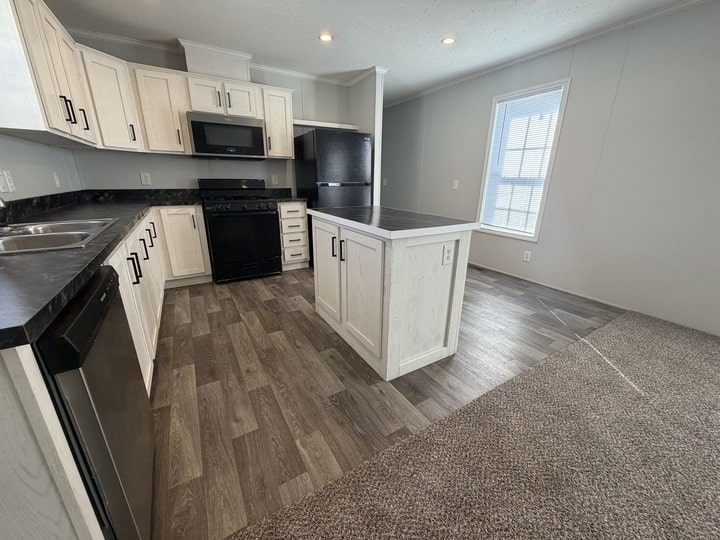 Modern kitchen with white cabinets, black countertops, stainless steel appliances, a small central island, and wood-look flooring. A window lets in natural light, and carpeted flooring transitions from the kitchen to a living area.