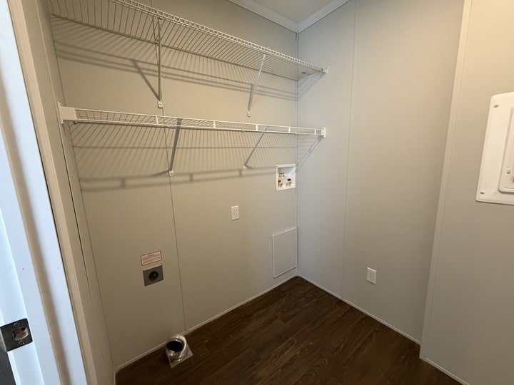 Empty laundry room with light-colored walls, two wire shelves, hookups for a washer and dryer, a small wall panel, and dark wood flooring. No appliances or other furniture are present.