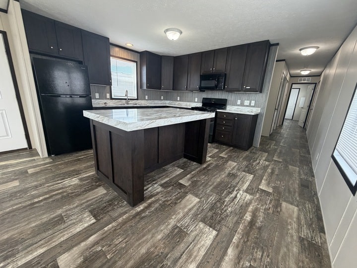 Modern kitchen with dark wood cabinets, a large island with a marble countertop, black appliances, wood-look flooring, and a hallway leading to other rooms. Natural light comes through a window above the sink.