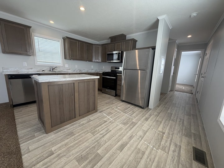 Modern kitchen with wood cabinets, stainless steel appliances, a center island, and light wood-style flooring. A hallway leads away from the kitchen, with natural light coming from windows.