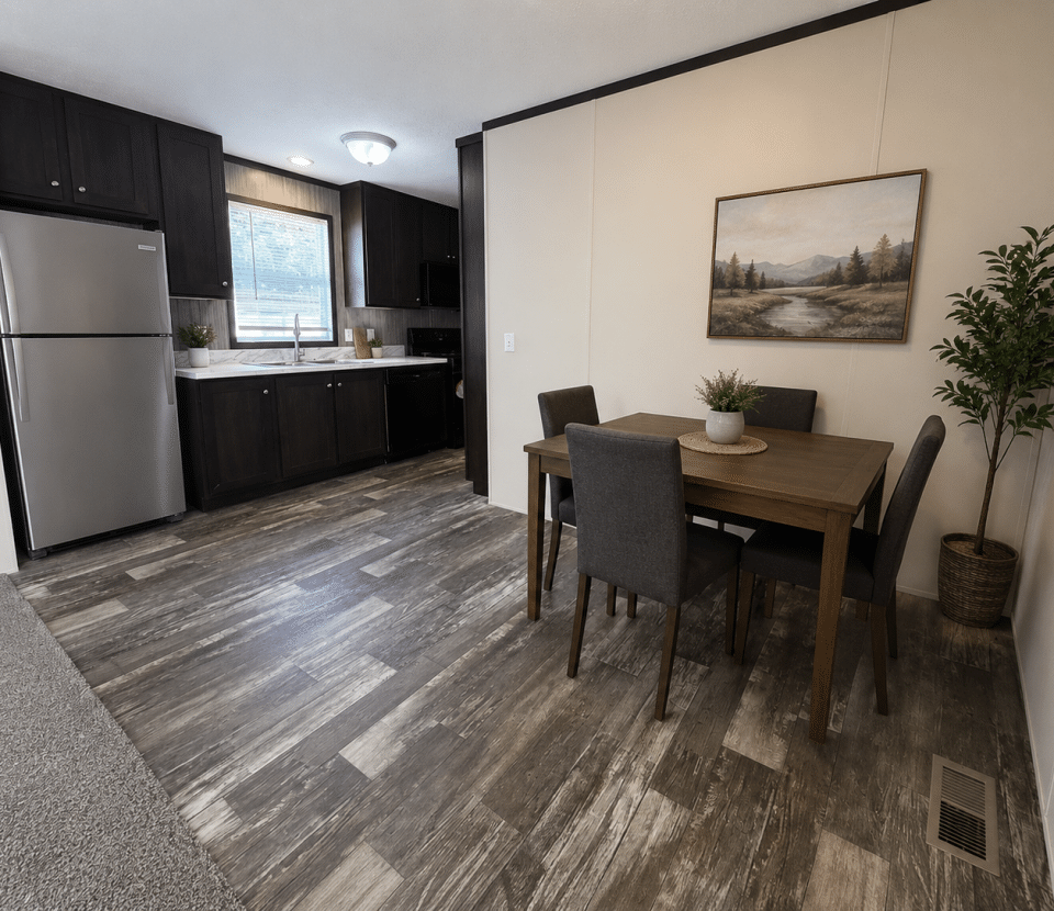 Modern kitchen and dining area with dark cabinets, stainless steel refrigerator, a wooden dining table with four chairs, a potted plant, and a landscape painting on a cream-colored wall.