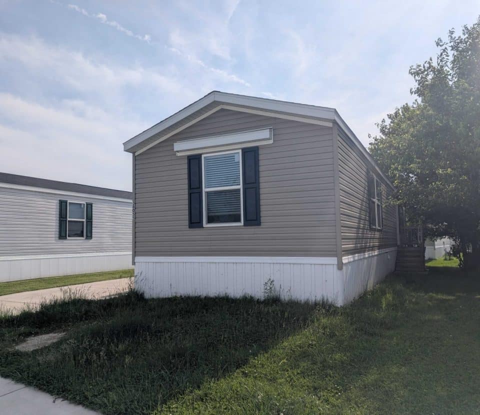 A beige manufactured home with white trim and dark shutters sits on a grassy lot, with another similar home visible in the background under a partly cloudy sky.