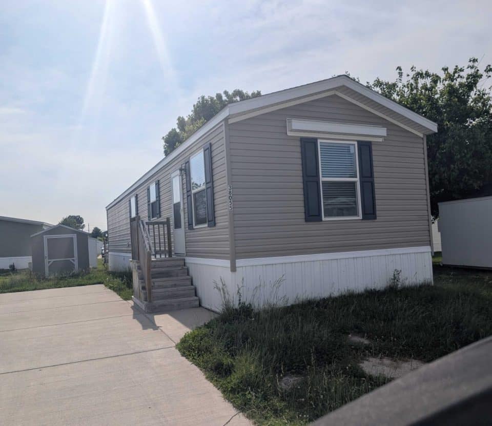 A beige mobile home with white trim and dark shutters sits on a concrete driveway, with steps leading to the entrance and some grass growing around the base. Another mobile home and shed are visible in the background.