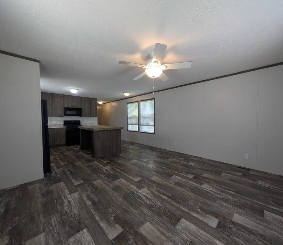 Spacious, empty living area with dark wood-style flooring, beige walls, large windows, a ceiling fan with lights, and an open kitchen featuring dark cabinets and appliances.