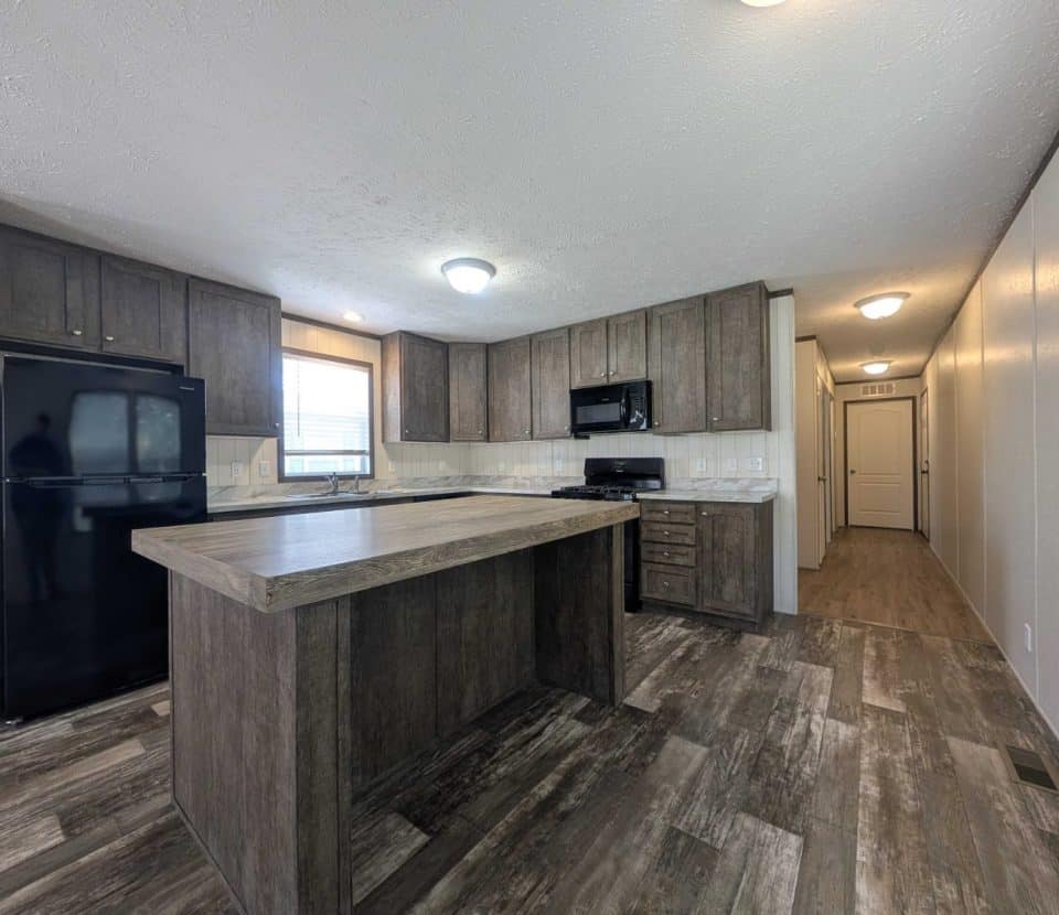 Spacious kitchen interior with dark wood cabinets, a large island, black refrigerator, black oven, and microwave. The room features wood-style flooring and neutral-colored walls, with natural light coming from a window.