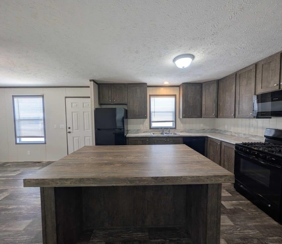 Modern kitchen with dark wood cabinets, black appliances, a large center island, and a tile backsplash. Natural light enters through two windows, and gray flooring extends throughout the room.