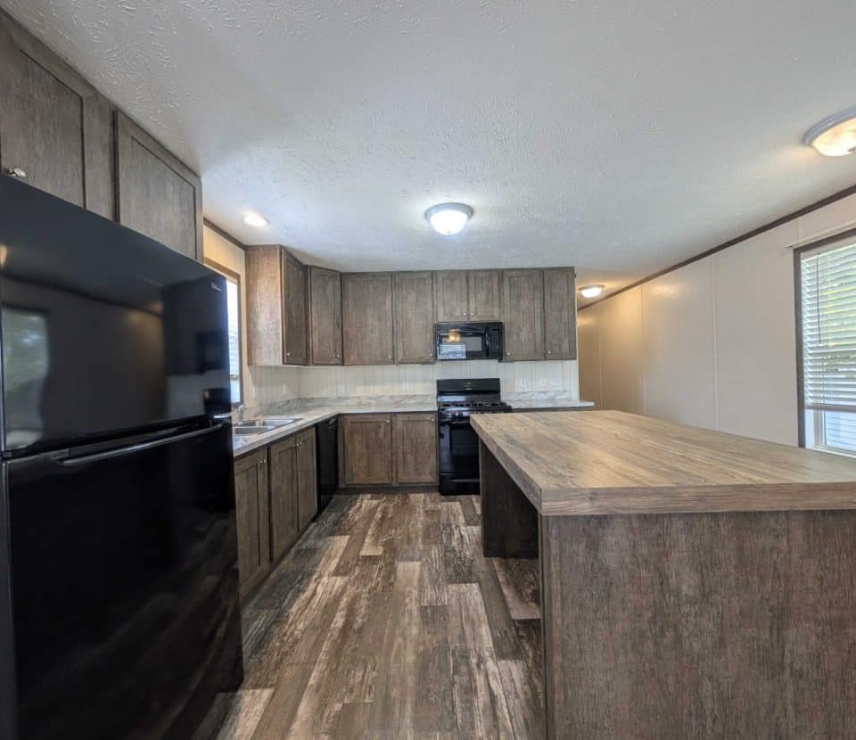 Modern kitchen with wood-look cabinets and flooring, black refrigerator, stove, and microwave, a large central island, and ample counter space. Two ceiling lights and a window provide bright illumination.