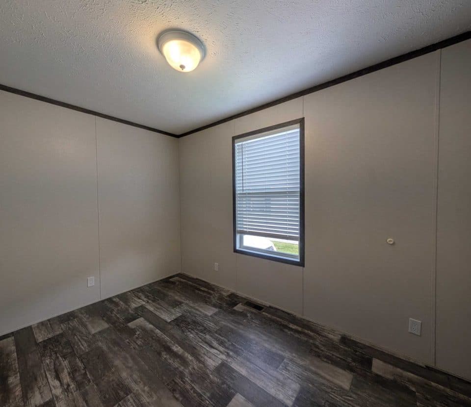 Empty room with beige walls, a single window with blinds, a ceiling light, and dark wood-patterned flooring. The space is unfurnished and well-lit by natural light from the window.