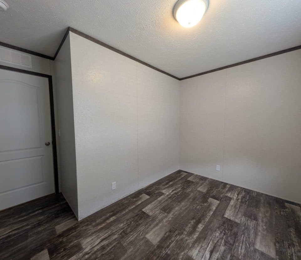 Empty room with plain beige walls, dark wood-style flooring, a white ceiling light, and a closed white door; the space is unfurnished and well-lit.