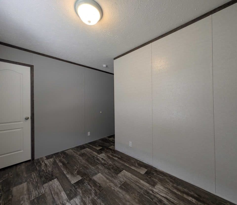 A small, empty room with gray and beige walls, a white ceiling light, a closed white door, dark wood-patterned flooring, and no furniture.