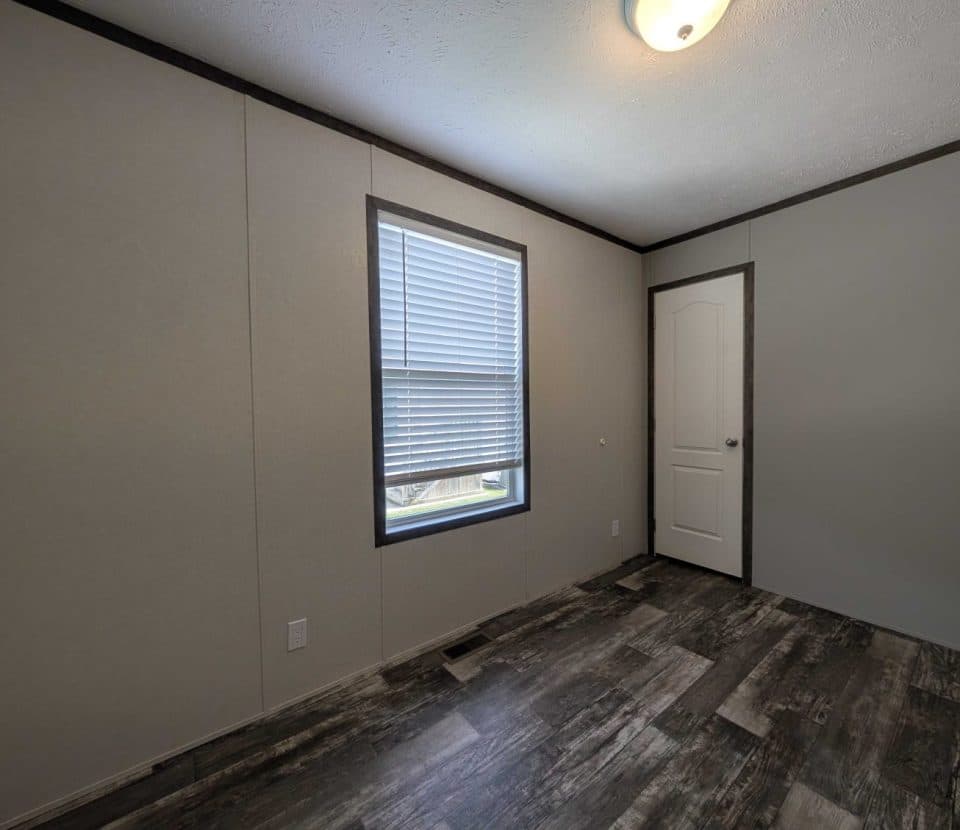 An empty room with light gray walls, a single window with closed blinds, dark trim, a white door, and wood-patterned vinyl flooring. A ceiling light fixture is visible near the corner.
