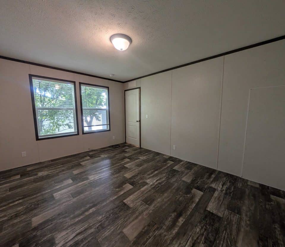 A mostly empty room with wood-patterned flooring, beige walls, a ceiling light, two large windows with a view of tree branches, and a closed door on the far wall.