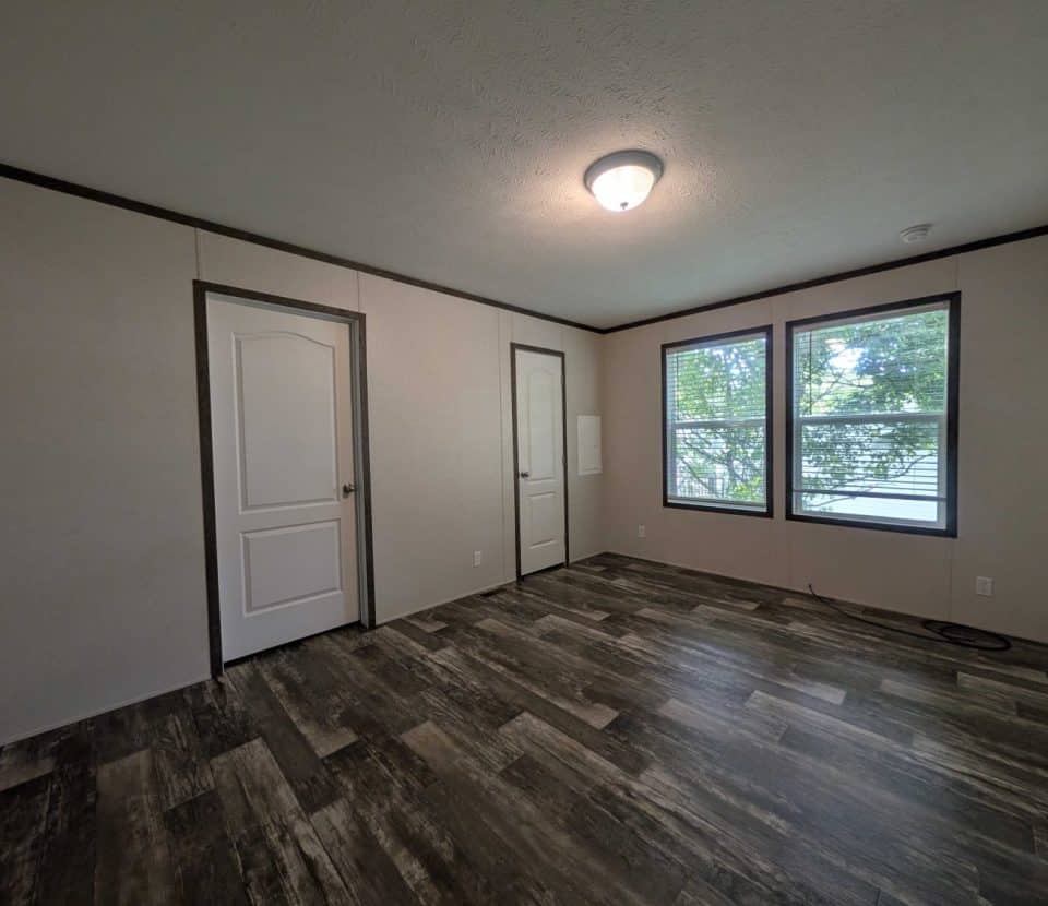 A bright, empty room with light-colored walls, dark trim, wood-patterned flooring, two white doors, and three large windows letting in natural light with trees visible outside.