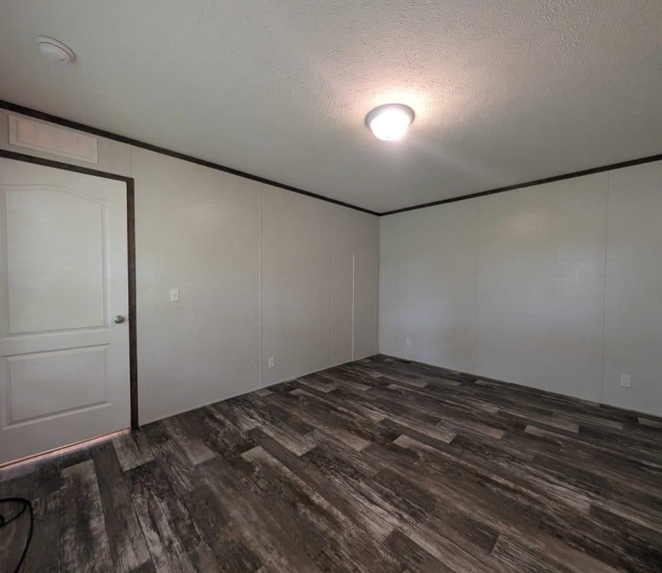 Empty room with light-colored walls, a closed white door, dark trim, wood-patterned flooring, a ceiling light, and electrical outlets along the walls. No furniture or decorations are present.