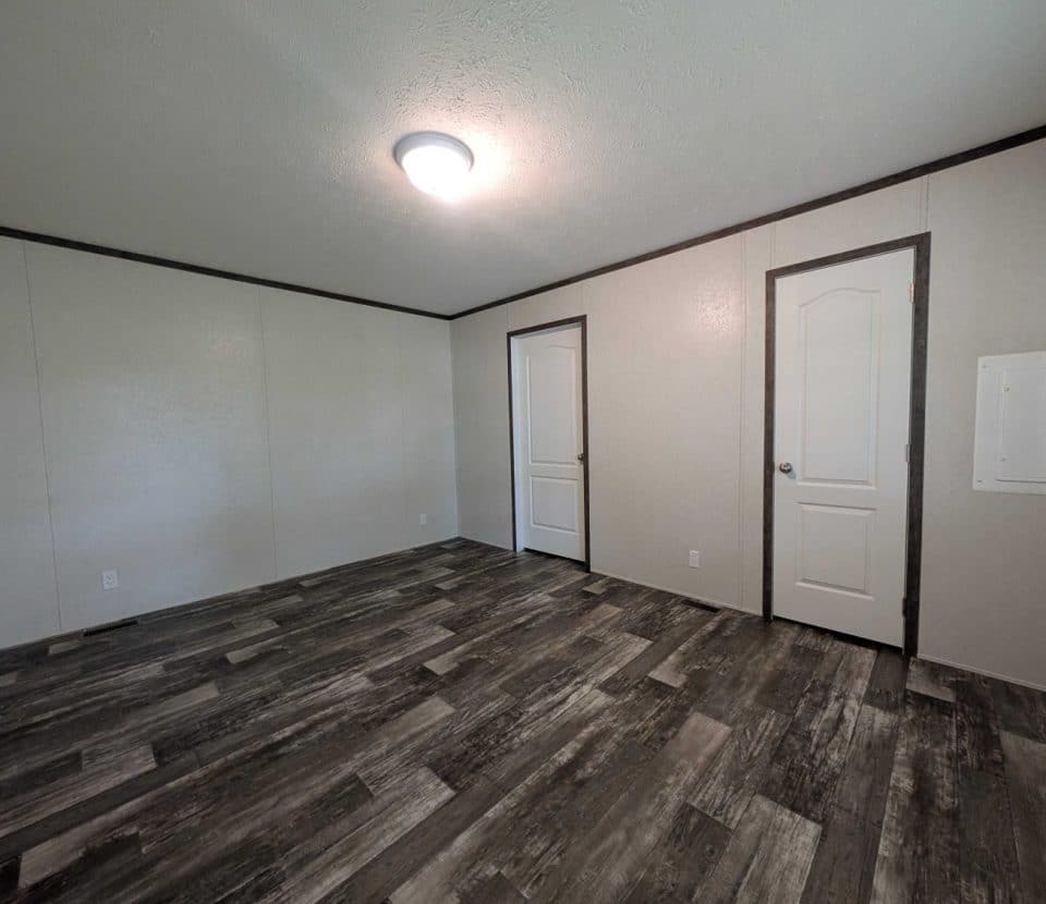 Empty room with light-colored walls, dark trim, two white doors, a ceiling light, and gray wood-textured flooring. There is also a small white electrical panel on the right wall.