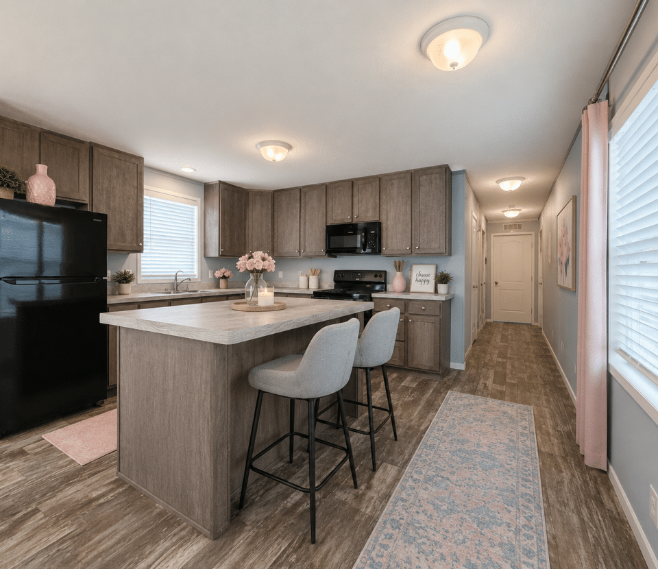 Modern kitchen with wood cabinets, black appliances, a center island with two gray stools, and light wood flooring. Pink accents and decor add warmth, while large windows let in natural light. A hallway leads to the back of the home.