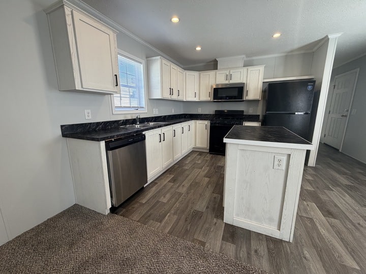 Modern kitchen with white cabinets, dark countertops, stainless steel dishwasher, black appliances, and an island. The space has wood-look flooring and neutral walls, with a window above the sink letting in natural light.