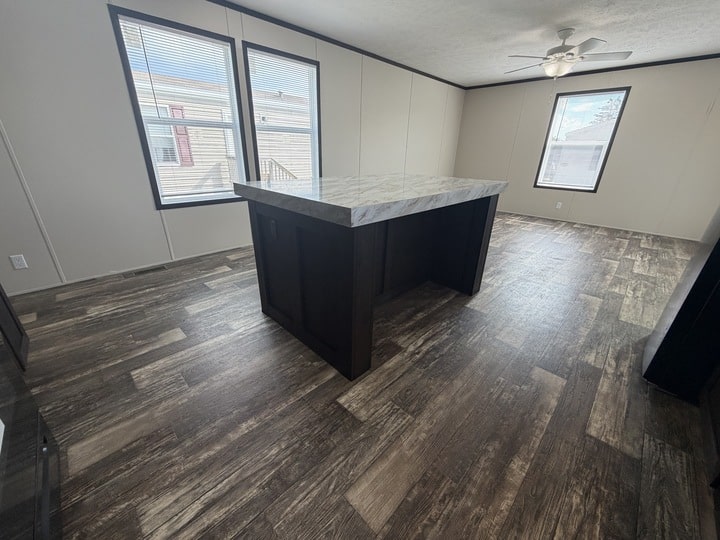 A spacious room with wood-patterned flooring, light-colored walls, large windows with blinds, a ceiling fan, and a central island with a marble countertop.