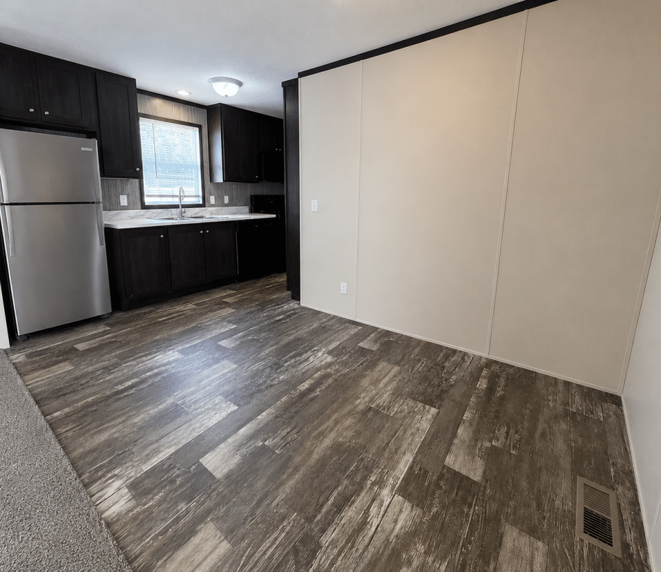 A modern kitchen with dark wood cabinets, stainless steel refrigerator, a window above the sink, and an adjacent empty area with wood-look flooring and beige walls.